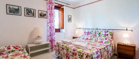 Un dormitorio con cama, mesita de noche, silla y ventana con cortinas.