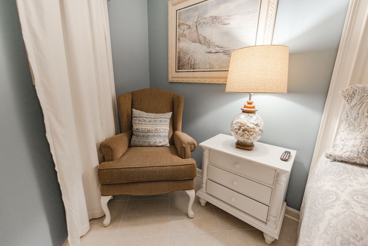 A room with a brown armchair, a white nightstand with a lamp, and a framed picture on the wall.
