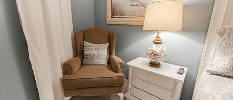 A room with a brown armchair, a white nightstand with a lamp, and a framed picture on the wall.