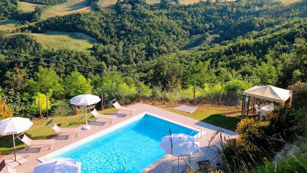 A pool area with lounge chairs and umbrellas, surrounded by lush greenery and hills in the background. - Villa in Marche With Pool & Panoramic Views (Acqualagna)