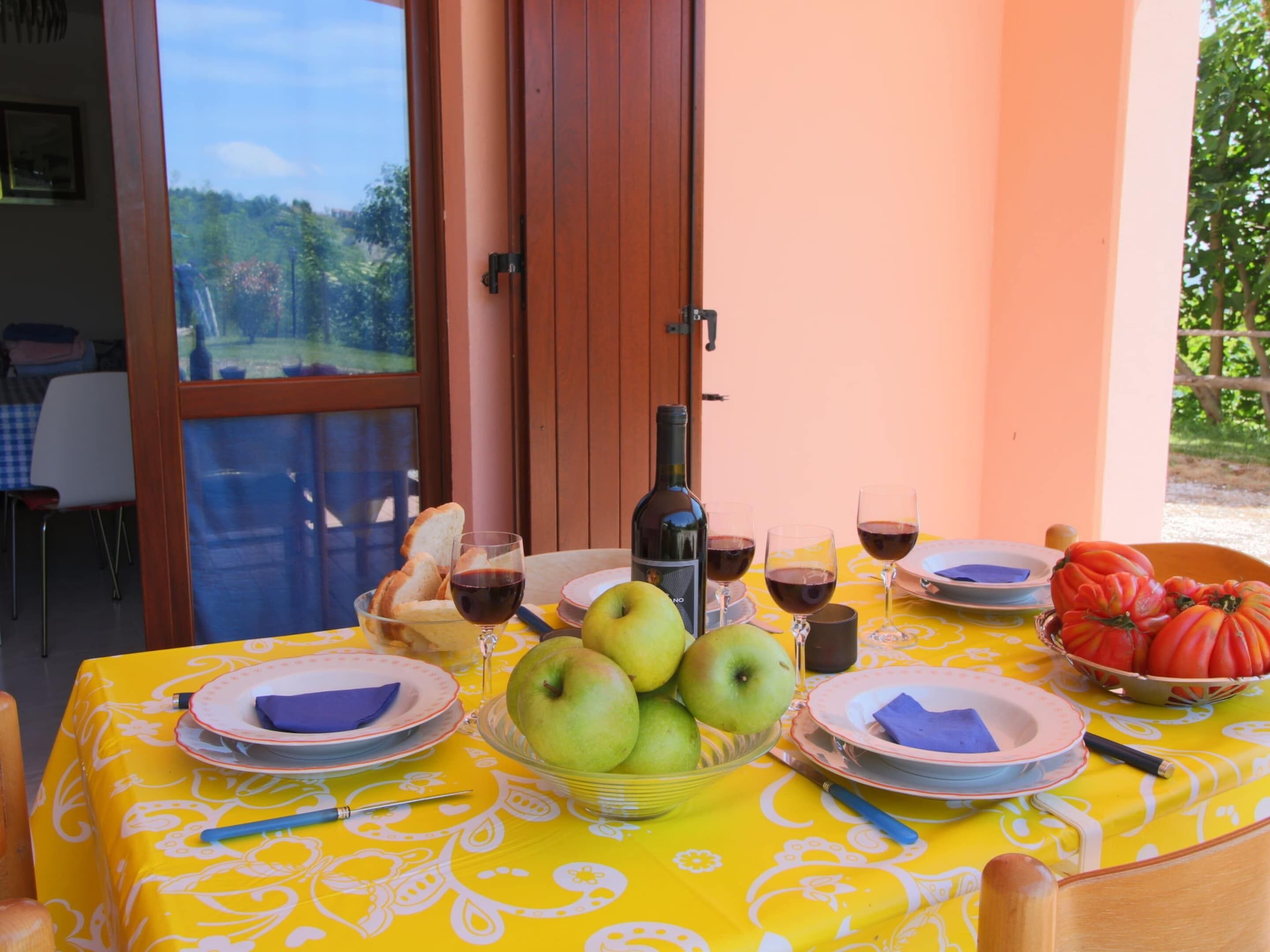 An outdoor dining area with a table set for two, featuring a bottle of wine, glasses, plates, a bowl of apples, and a basket of tomatoes.
