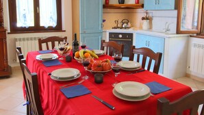 A dining area with a red tablecloth, set for a meal with plates, glasses, and a bottle of wine. There are chairs around the table, and a kitchen area with blue cabinets is visible in the background. - Villa in Marche With Pool & Panoramic Views (Acqualagna)