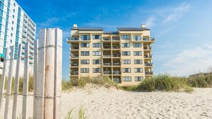 A beachfront hotel with multiple floors, balconies, and a sandy beach in the foreground.