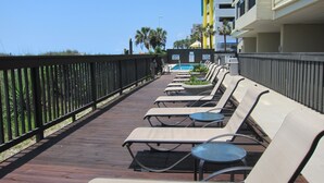 A wooden deck with lounge chairs and tables, a pool, and a building with balconies.