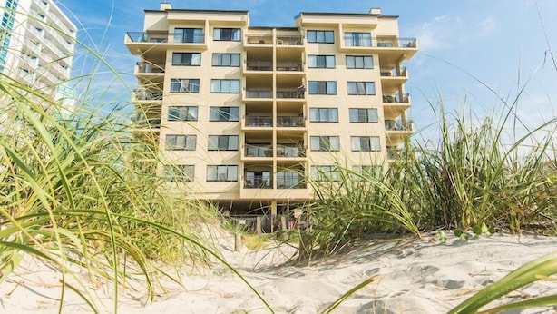 Ein Hotel direkt am Strand mit mehreren Etagen, umgeben von Sand und Strandgras.