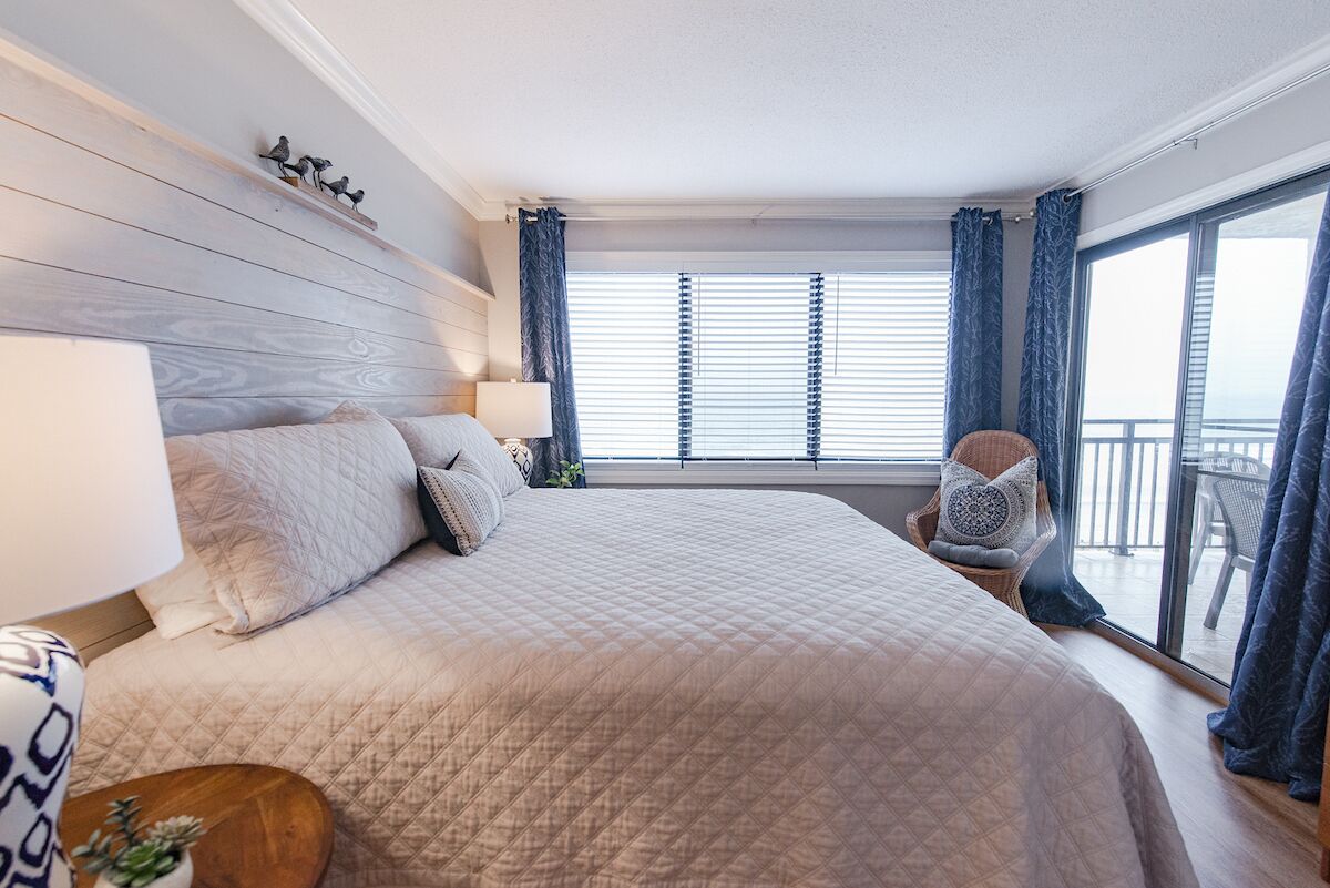 Un dormitorio con una cama grande, una mesita de noche, una silla y un ventanal con cortinas.