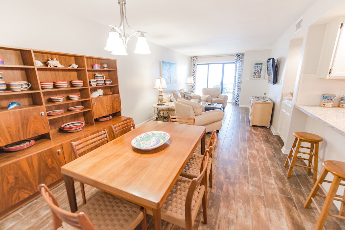 A spacious kitchen with wooden cabinets, a dining table, and chairs. There is a kitchen island with a lamp, and a living area with a sofa and a coffee table.