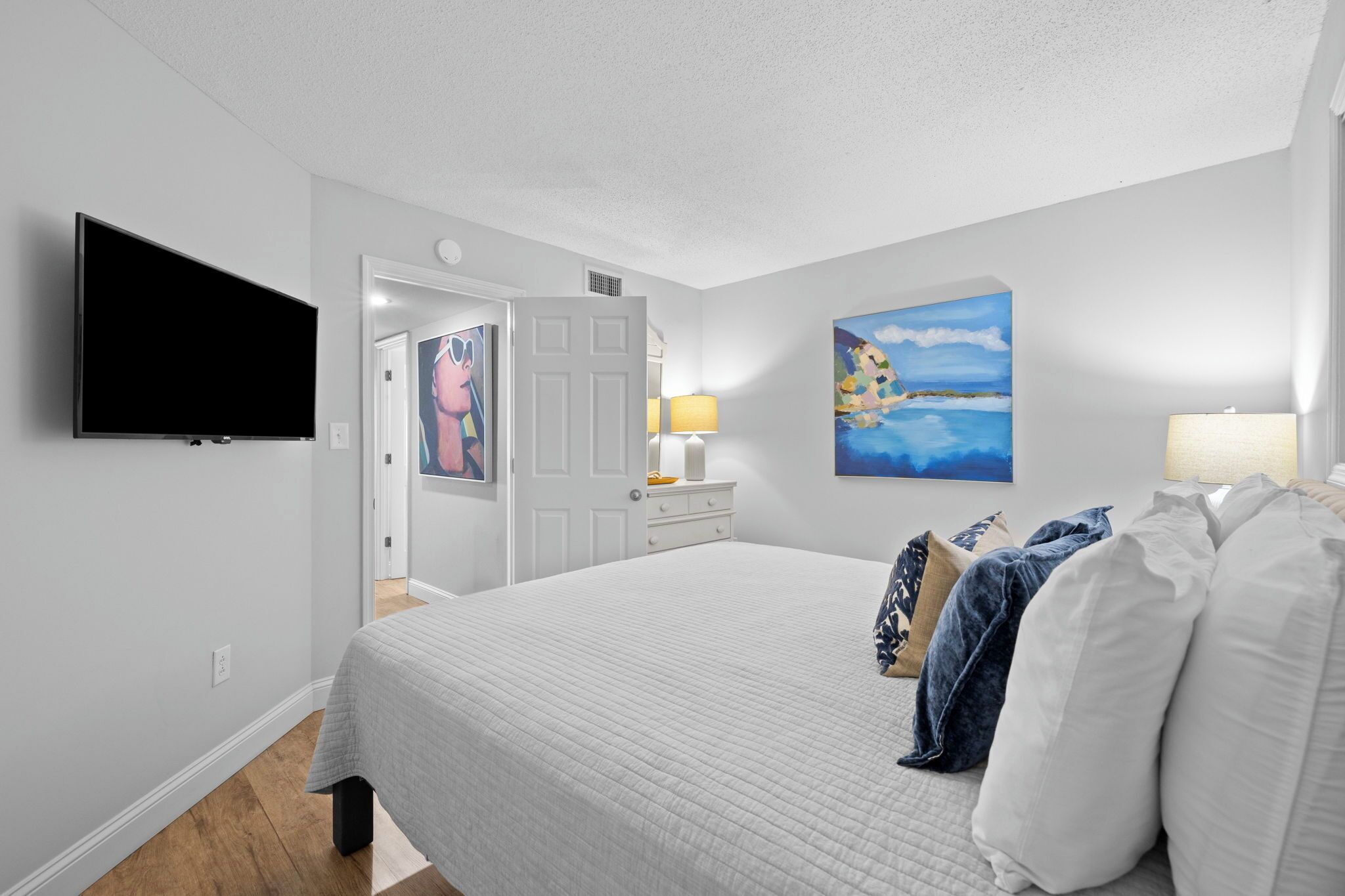 Un dormitorio con una cama grande, un televisor de pantalla plana montado en la pared, una puerta que da a otra habitación y un cuadro en la pared.