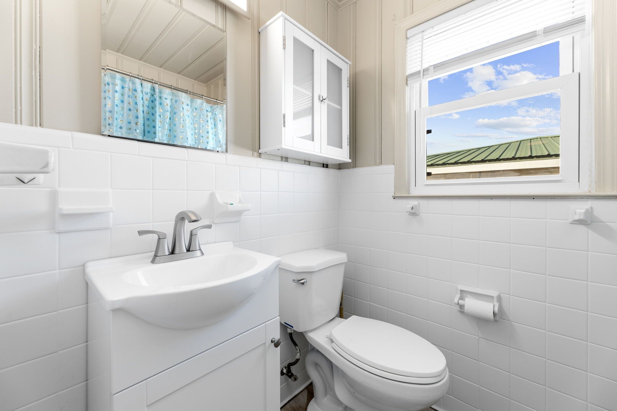 Une salle de bain avec un lavabo, des toilettes et un meuble de rangement, tous de couleur blanche, une fenêtre donnant sur un bâtiment doté d’un toit végétalisé, et un rideau de douche bleu.