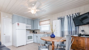 Une cuisine compacte avec un réfrigérateur, une cuisinière et un four à micro-ondes blancs, une table à manger avec quatre chaises, un ventilateur de plafond et un téléviseur posé sur une commode en bois.