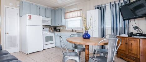 A compact kitchen with a white refrigerator, stove, and microwave, a dining table with four chairs, a ceiling fan, and a television on a wooden cabinet.