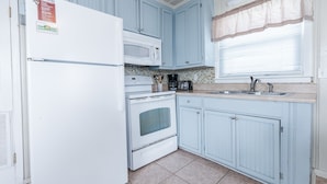 Une cuisine avec des meubles de couleur bleu clair, un réfrigérateur blanc, un four à micro-ondes, une cuisinière et une fenêtre ornée d’un rideau beige.