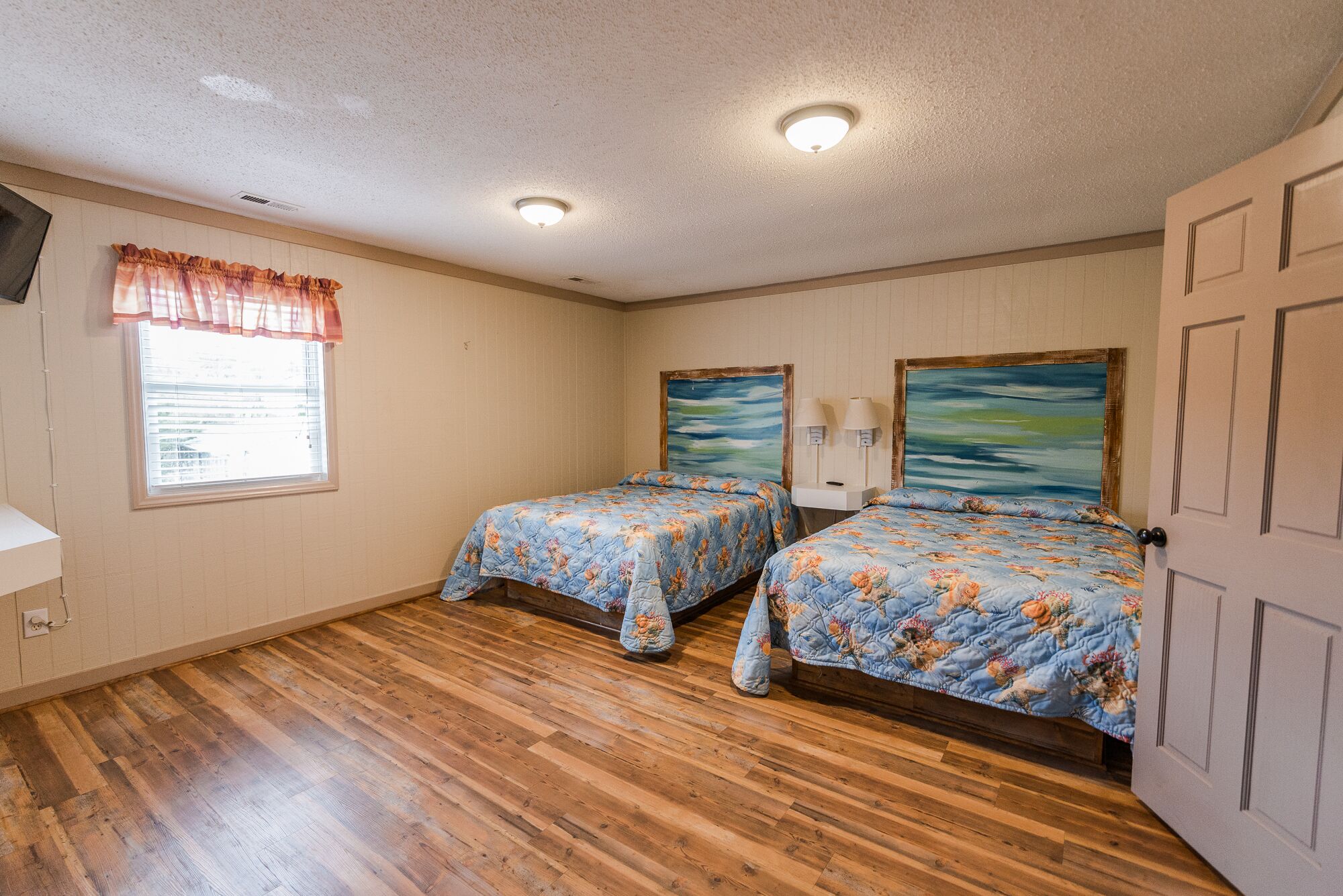 A bedroom with two beds, wooden flooring, a window with blinds, and a door.