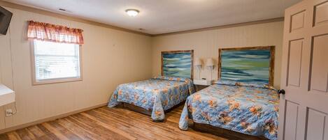 A bedroom with two beds, wooden flooring, a window with blinds, and a door.