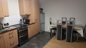 Een kleine keuken met houten kastjes, een gasfornuis, een oven, een koelkast en een eettafel met stoelen.