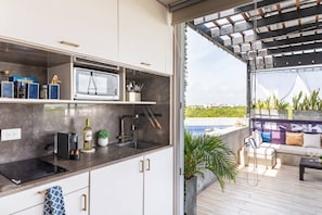 Cozinha moderna com micro-ondas embutido, pia e bancada com diversos objetos. Há uma porta deslizante que dá para um pátio externo com piscina e área de estar.