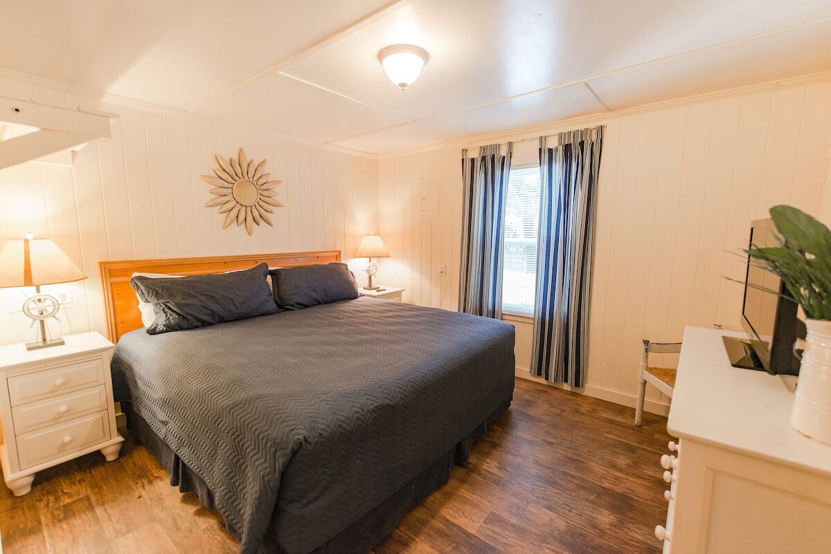 A bedroom with a large bed, two bedside tables with lamps, a wooden headboard, a sun-shaped wall decoration, a window with curtains, and a small chair.