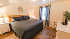 Une chambre à coucher avec un grand lit, deux tables de chevet avec des lampes, une tête de lit en bois, une décoration murale en forme de soleil, une fenêtre avec des rideaux et une petite chaise.