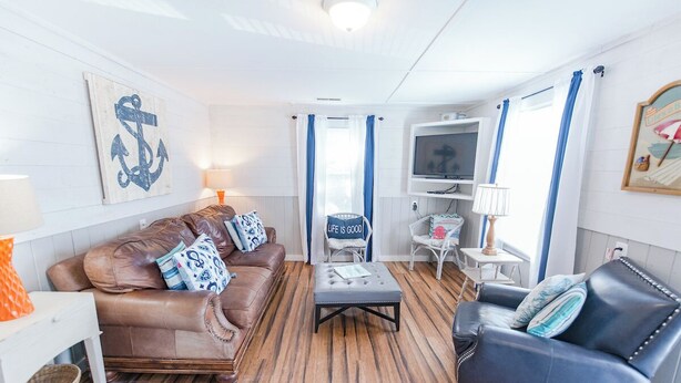 Un salon chaleureux avec un canapé en cuir marron, un fauteuil bleu, un plancher en bois et un téléviseur posé sur un meuble blanc.