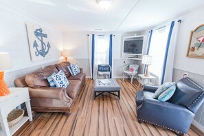 Una sala de estar acogedora con un sofá de cuero marrón, un sillón azul, piso de madera y un televisor sobre un soporte blanco.