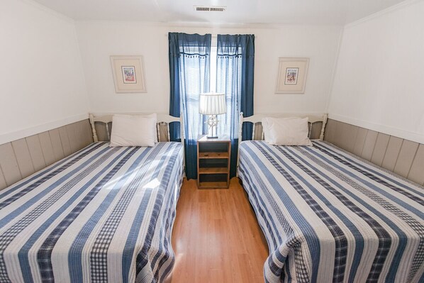 Habitación doble con dos camas individuales, suelo de madera, mesita de noche con lámpara y ventana con cortinas.