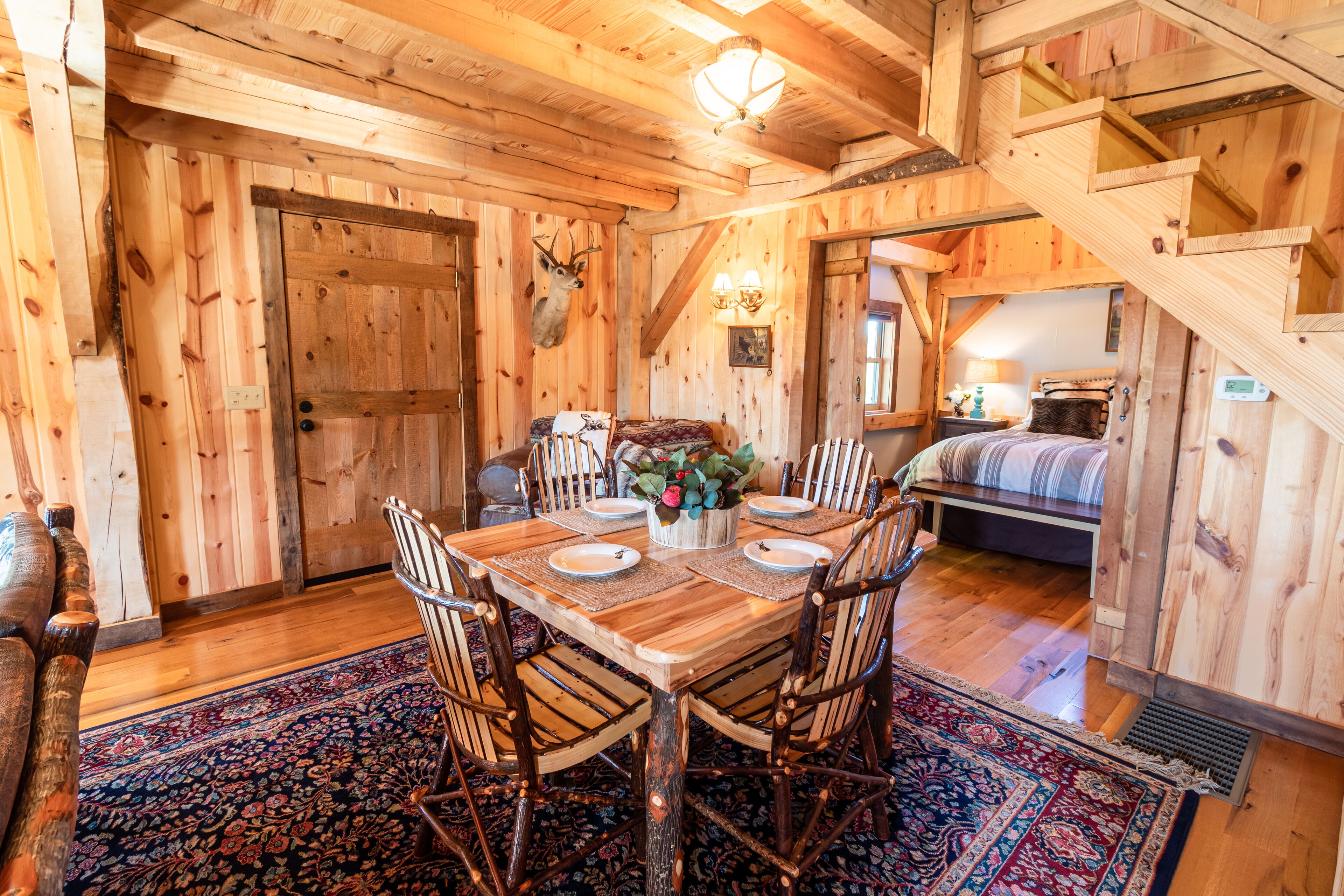A rustic wooden dining room with a large table set for six, surrounded by wooden chairs. A deer head is mounted on the wall above the door.