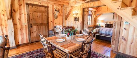 A rustic wooden dining room with a large table set for six, surrounded by wooden chairs. A deer head is mounted on the wall above the door.