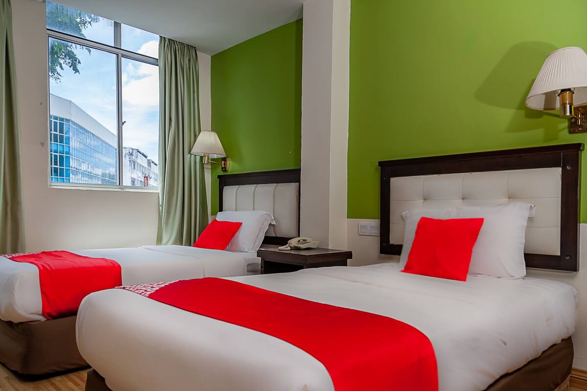 A hotel room with two beds, green walls, a window with a view of buildings, and a nightstand with a lamp.