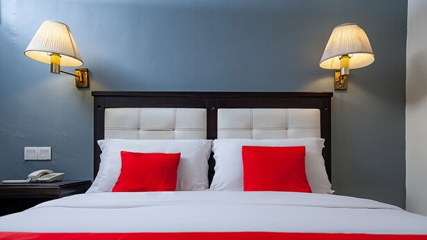 Un lit avec une literie blanche et rouge, deux lampes fixées au mur, un téléphone sur la table de chevet et une tête de lit sombre.