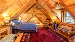 Een gezellig slaapkamer onder het dak, met houten balkenplafond, een groot bed, een rood kamerkleed met patroon en een bankje.