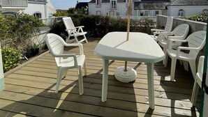 Eine Holzterrasse mit Tisch und Stühlen unter einem großen Sonnenschirm, umgeben von weißen Gebäuden.