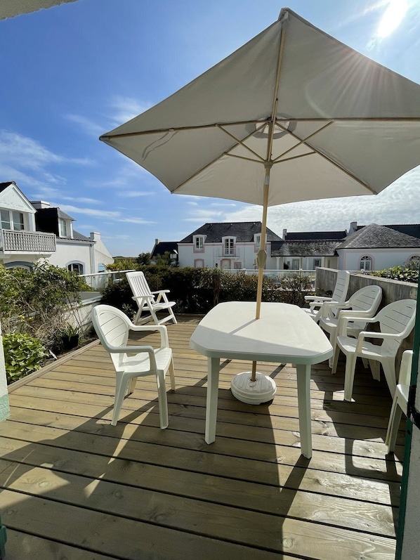 A wooden deck with a table and chairs under a large umbrella, surrounded by white buildings.