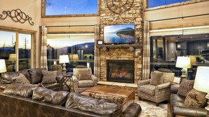 A spacious living room with leather sofas, patterned armchairs, a stone fireplace, and large windows offering scenic views.