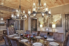 A dining area with a wooden ceiling, a chandelier, and a table set for six with wine glasses and plates.