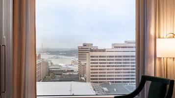 A hotel room with a view of a cityscape through a large window, a chair, and a lamp on the wall.