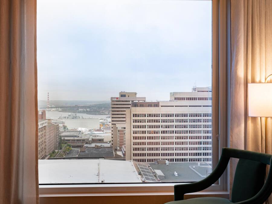 A hotel room with a view of a cityscape through a large window, a chair, and a lamp on the wall.