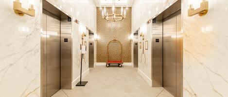 A modern elevator lobby with marble walls, a geometric patterned wall, and a central chandelier.