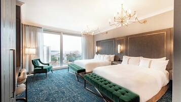 A hotel room with two beds, a green armchair, a desk, and a large window with a city view.