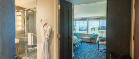 A hotel room with a glass-enclosed shower, a robe hanging on a hook, a bed with a blue carpet, and a seating area with a sofa and chairs.