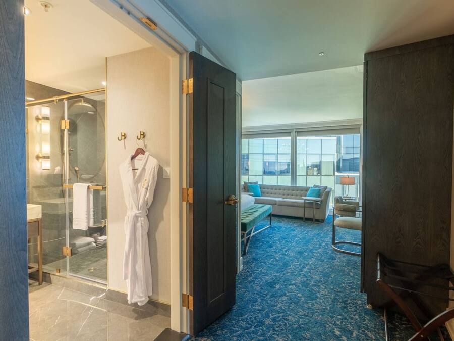 A hotel room with a glass-enclosed shower, a robe hanging on a hook, a bed with a blue carpet, and a seating area with a sofa and chairs.