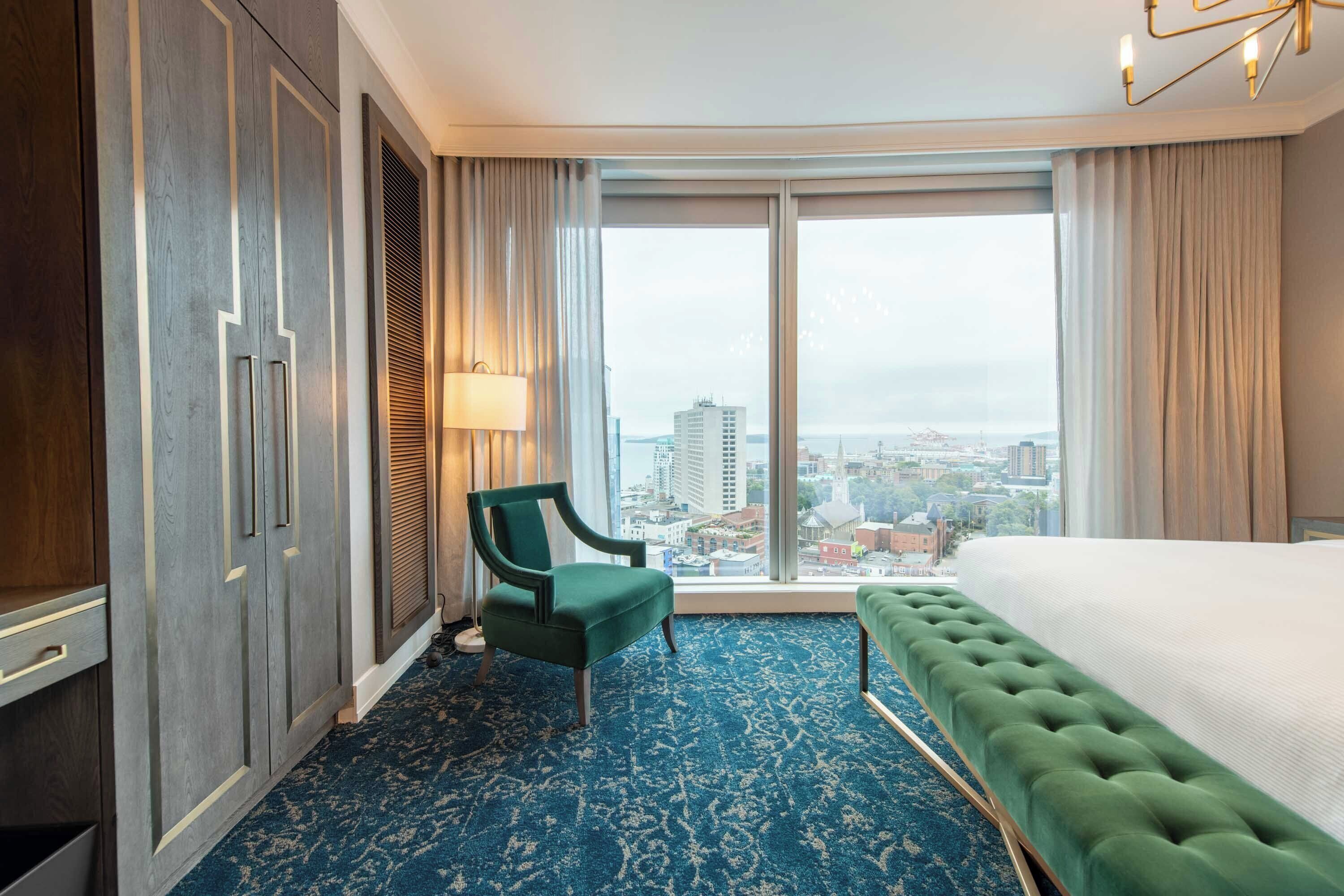 A hotel room with a large window, a green chair, a bed, and a blue carpet.