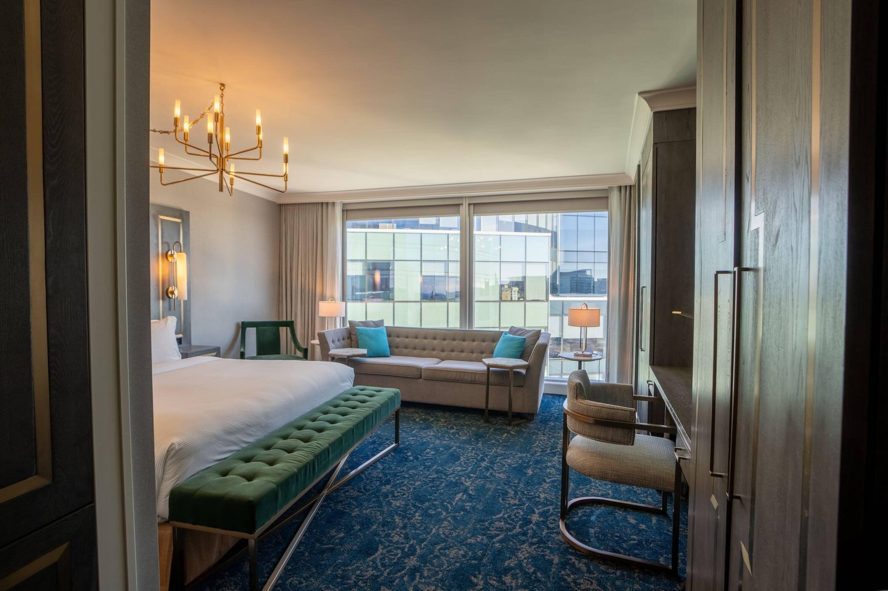 Une chambre d’hôtel avec un grand lit, un banc vert, un canapé et une chaise. On y trouve un lustre, une fenêtre donnant sur la ville et une porte.