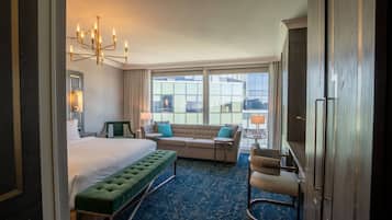 Une chambre d’hôtel avec un grand lit, un banc vert, un canapé et une chaise. On y trouve un lustre, une fenêtre donnant sur la ville et une porte.