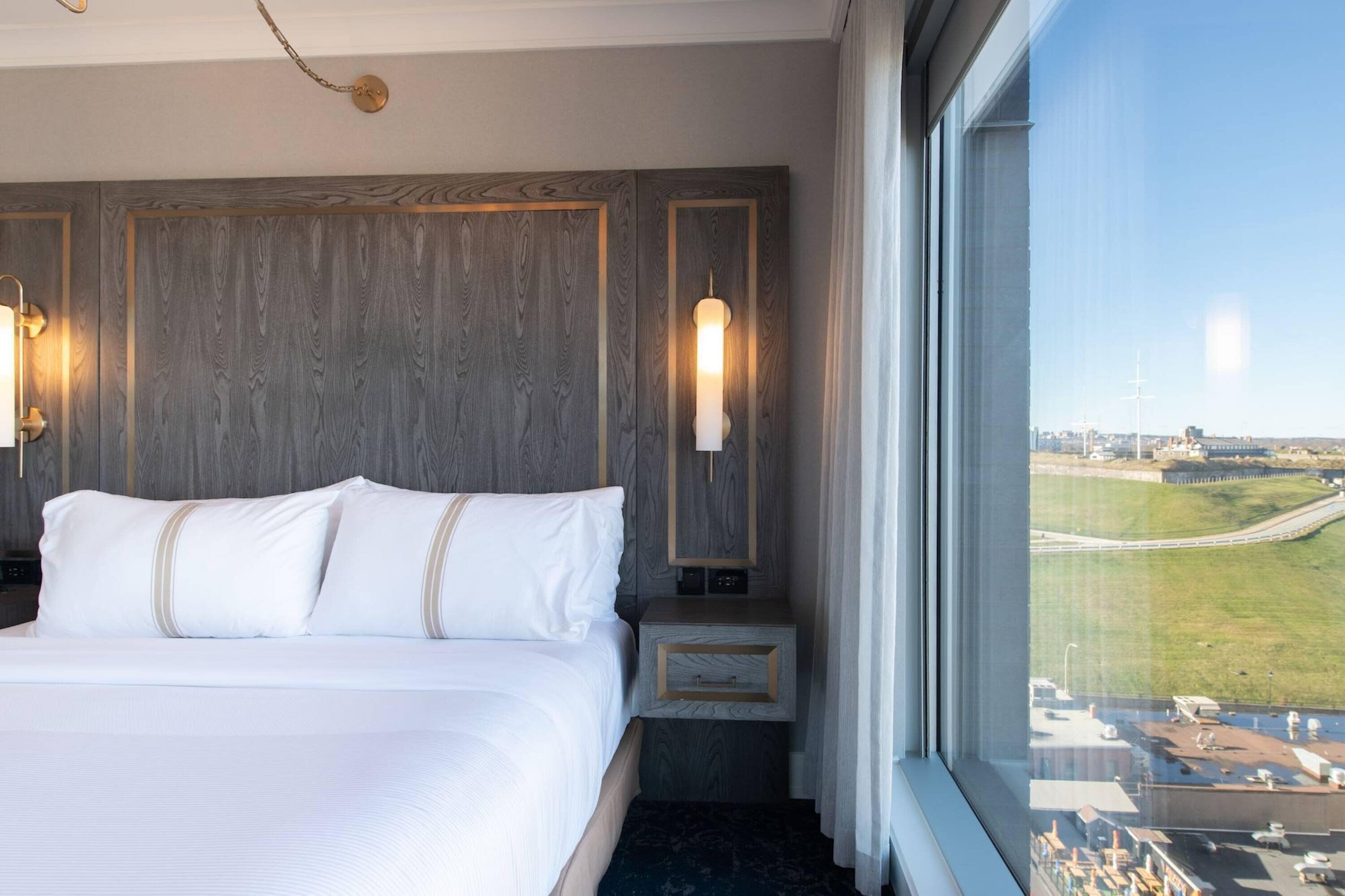 Une chambre d’hôtel avec un grand lit, des boiseries, deux lampes fixées au mur, une table de chevet et une fenêtre donnant sur un paysage urbain et un aéroport.
