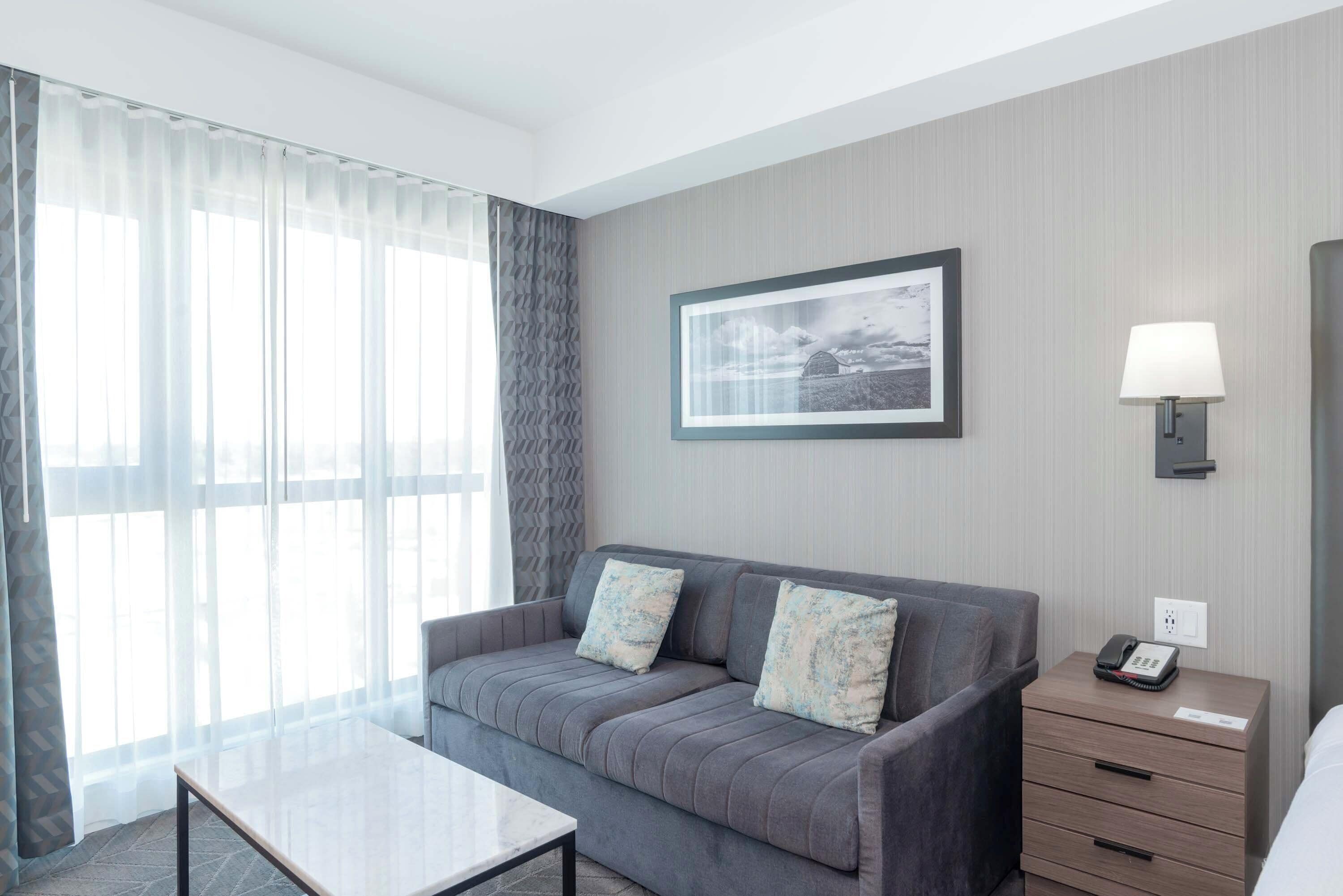 A hotel room with a grey sofa, a small table, a lamp, and a framed picture on the wall.