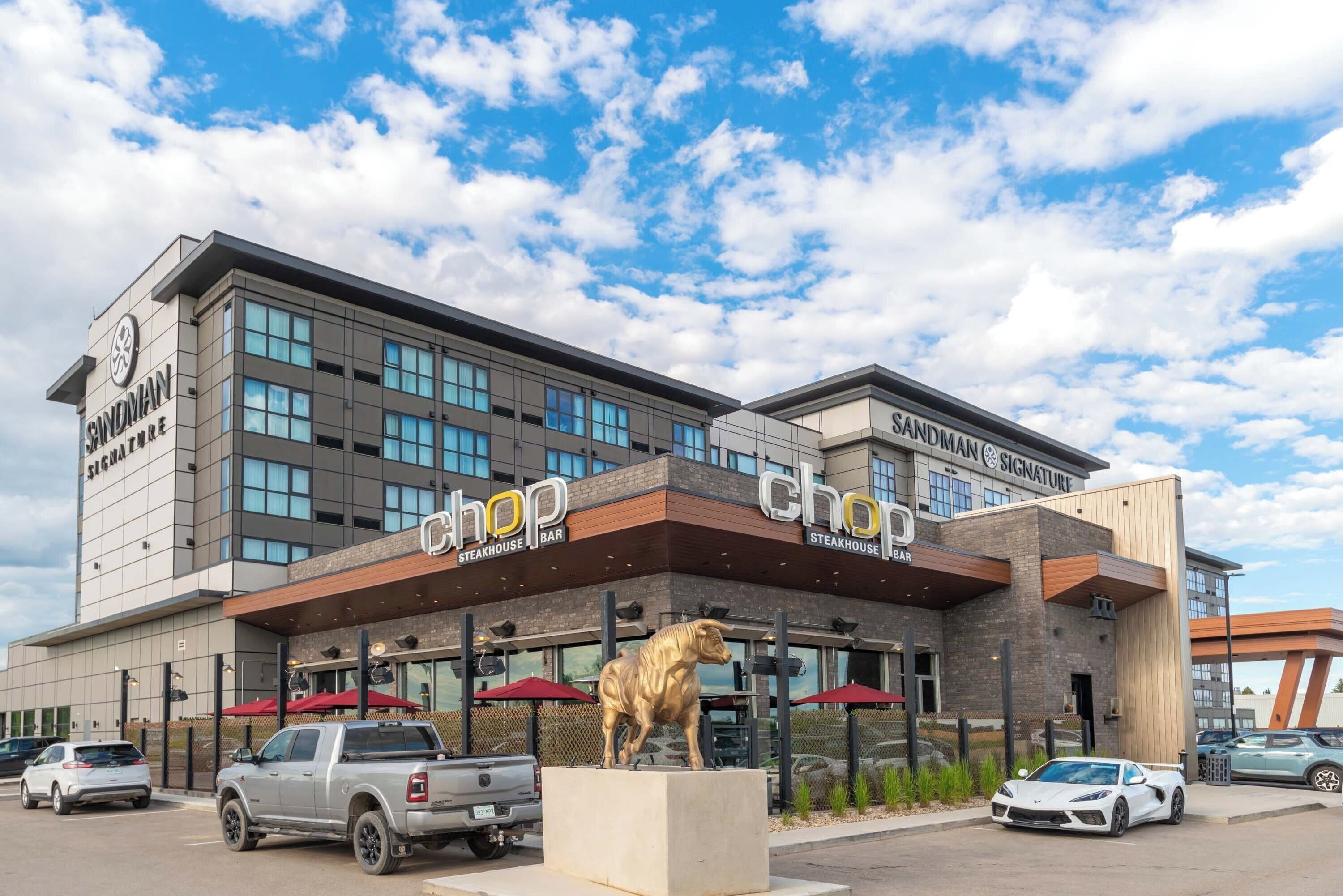 Un bâtiment moderne, sur lequel on peut lire l’enseigne « Sandman Signature Steakhouse », avec une grande statue de taureau doré devant et un parking où sont garées plusieurs voitures.