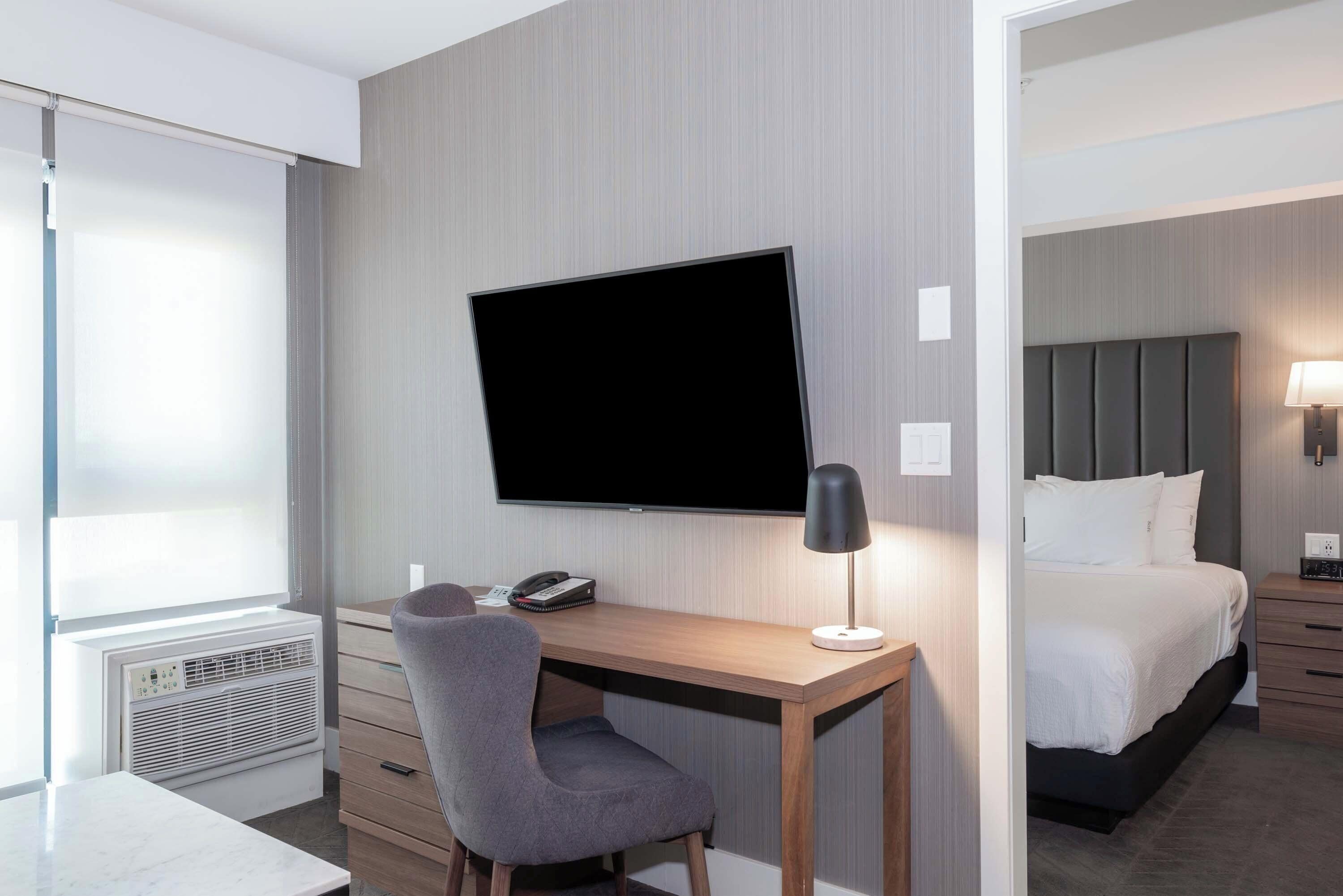 Camera d'albergo con una scrivania in legno, una televisione a schermo piatto, una poltrona grigia, un comodino con una lampada, un letto con biancheria bianca e una finestra con persiane.