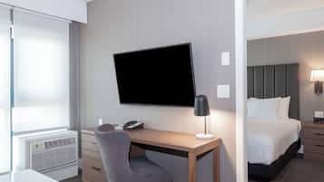 Camera d'albergo con una scrivania in legno, una televisione a schermo piatto, una poltrona grigia, un comodino con una lampada, un letto con biancheria bianca e una finestra con persiane.