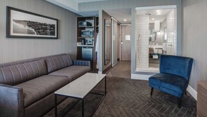 A modern hotel room with a sofa, a small table, a blue chair, a kitchenette, and a bathroom visible through an open door. - Sandman Signature Saskatoon South Hotel (Saskatoon)
