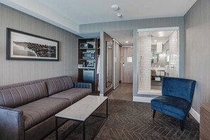 A modern hotel room with a sofa, a small table, a blue chair, a kitchenette, and a bathroom visible through an open door. - Sandman Signature Saskatoon South Hotel (Saskatoon)